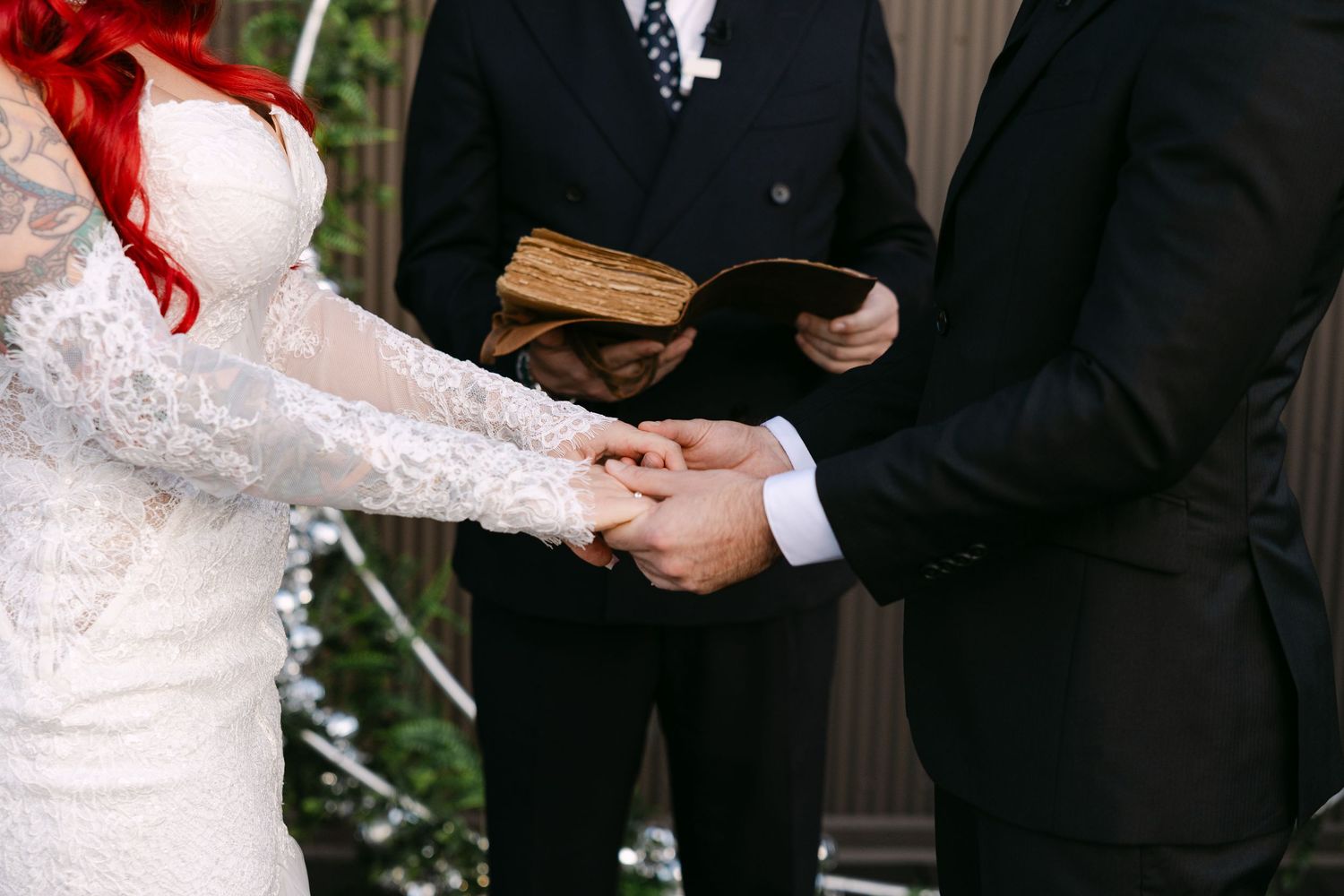 A wedding party stands in a line outdoors with greenery and a dark wall behind them.
