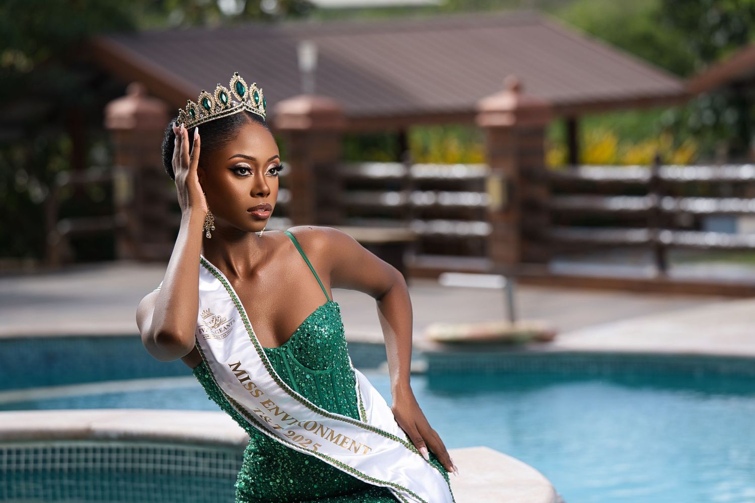 Beauty pageant contestant in a sparkling green gown and crown poses elegantly by a swimming pool.