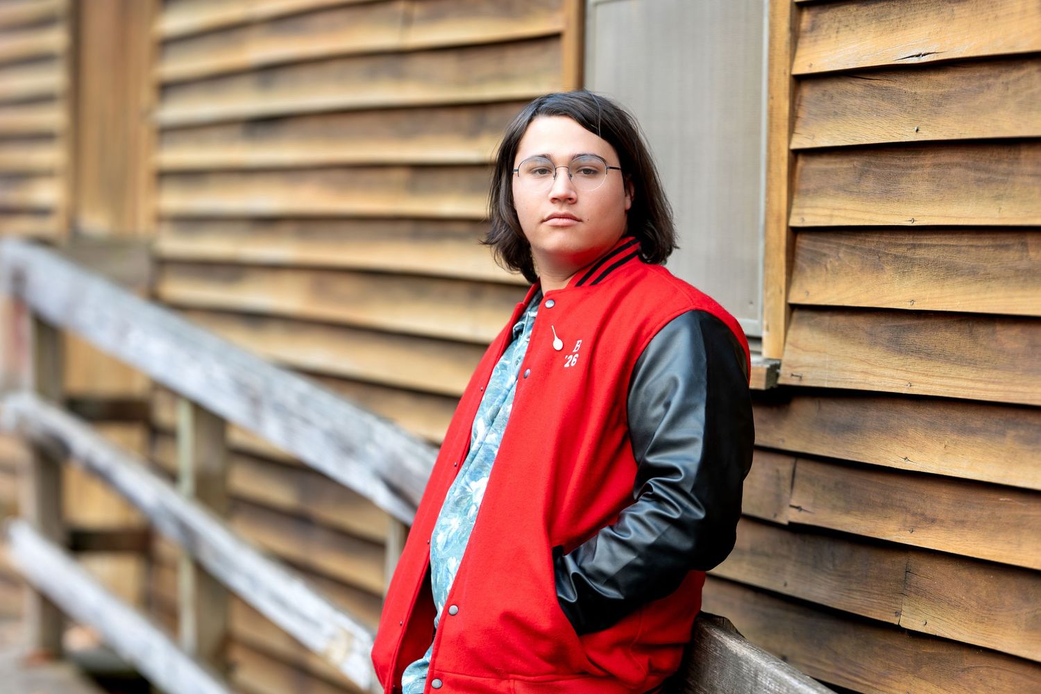 Young person in red varsity jacket with black leather sleeves leaning against wooden wall.