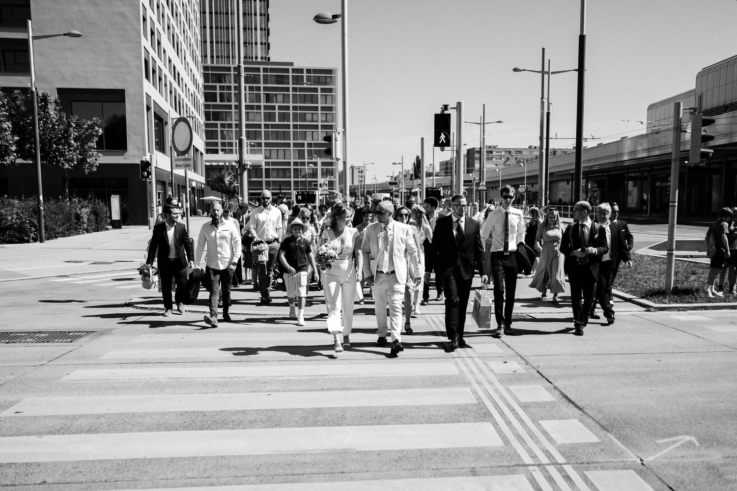 Black and white street scene showing people walking along an urban sidewalk on a sunny day.