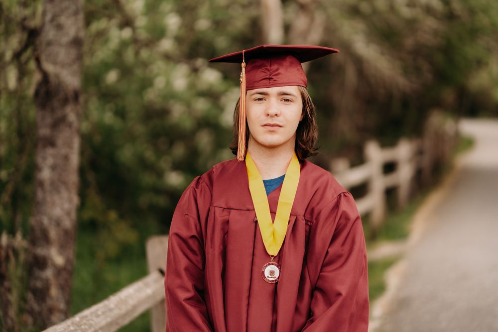 Graduate in burgundy cap and gown stands on tree-lined path during graduation photo session in Tacoma.