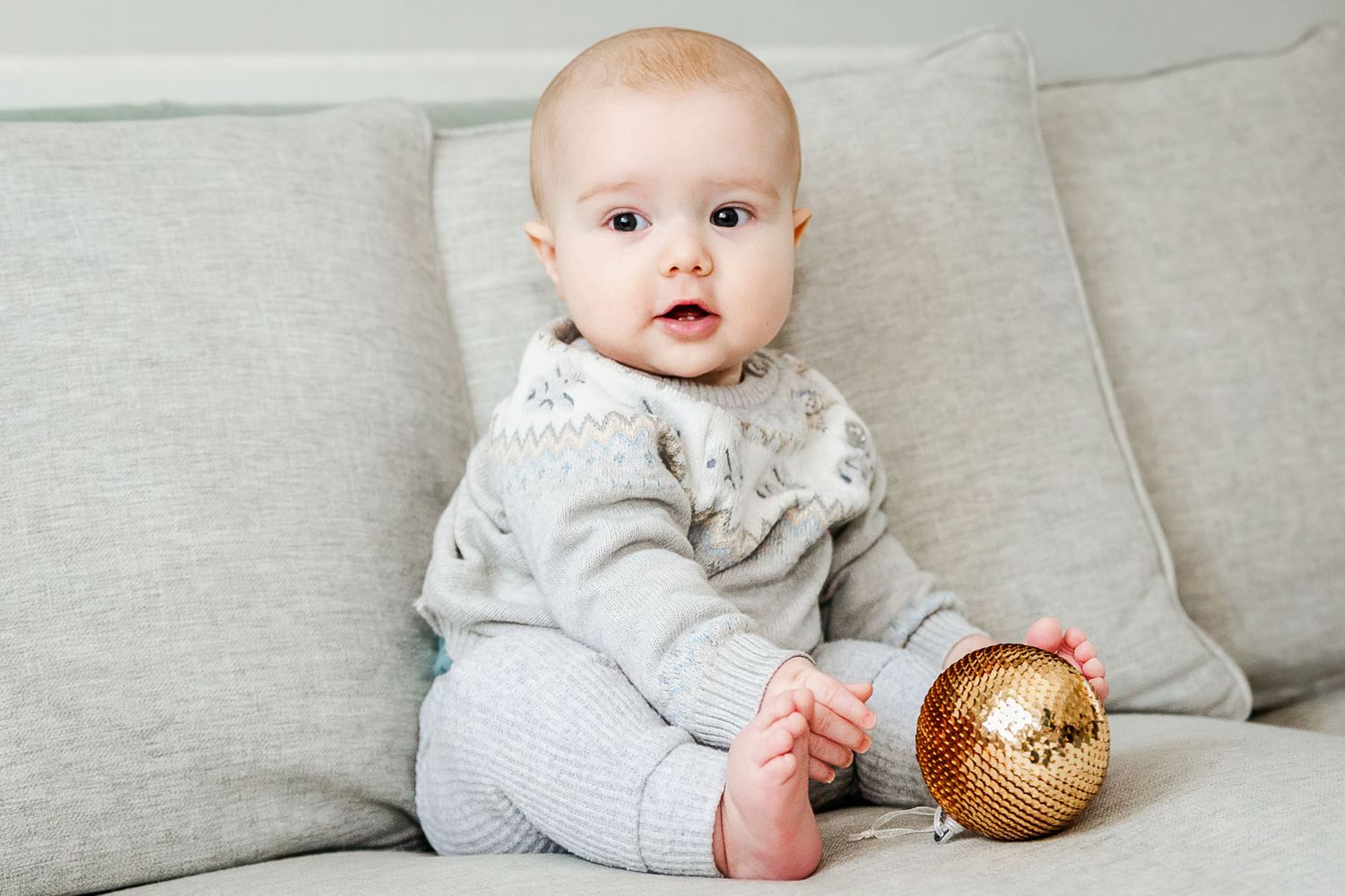 A baby looks happy as he sits on the sofa at home and plays with a bauble at a family Christmas photoshoot in Bristol