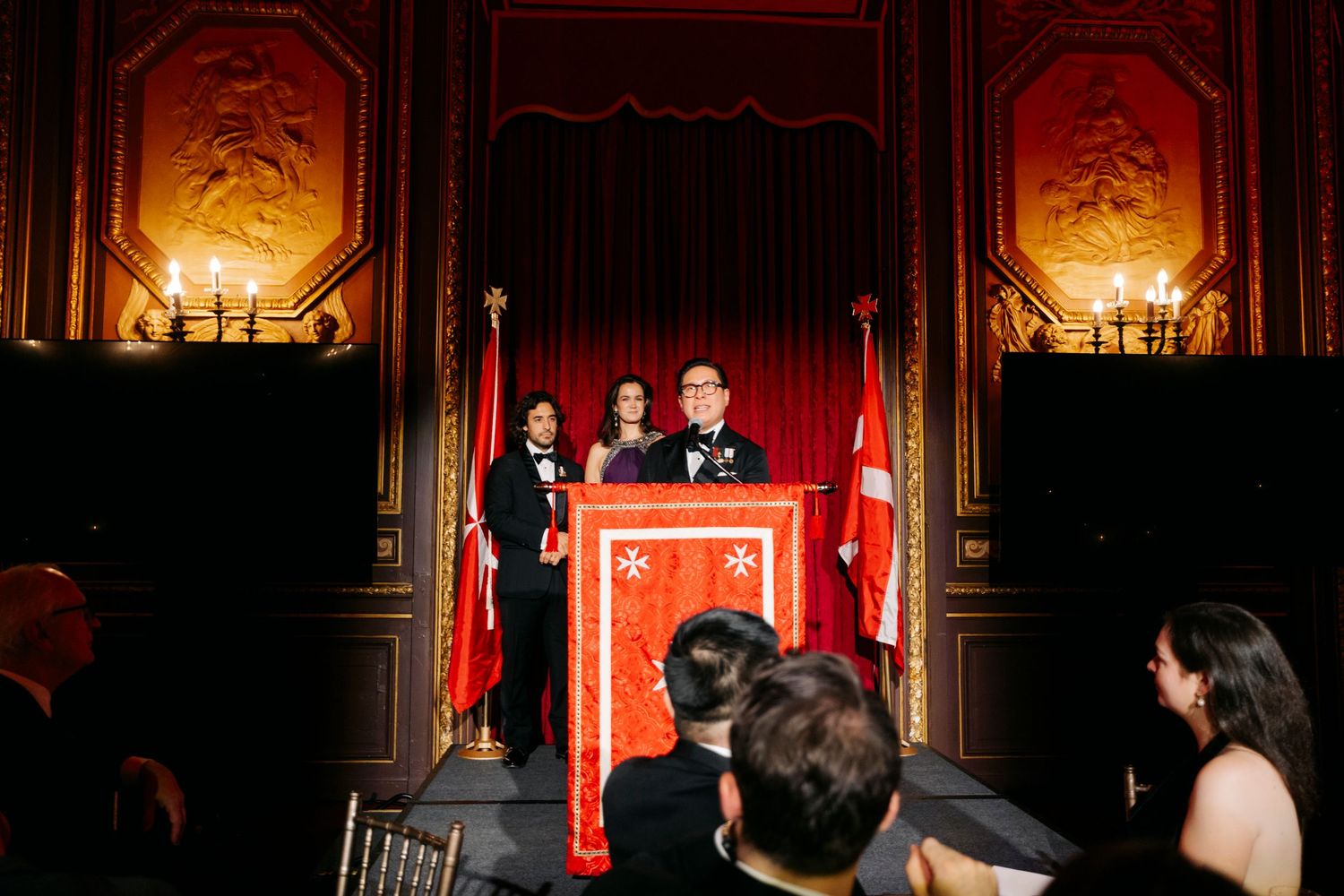 A dimly lit grand ballroom with ornate gold decorations and chandeliers casting warm light on red walls.