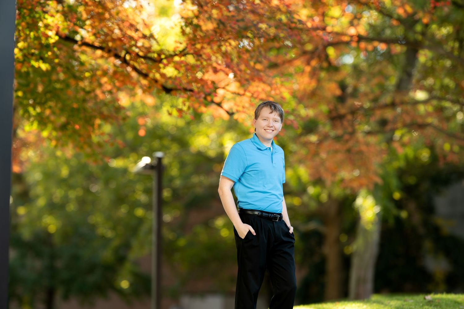 Young Asian man in blue polo shirt standing under vibrant red and orange autumn trees in a park.