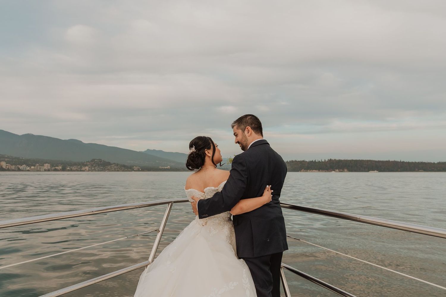Bride and groom embracing on the bow of a boat with a scenic lake and mountains behind them.
