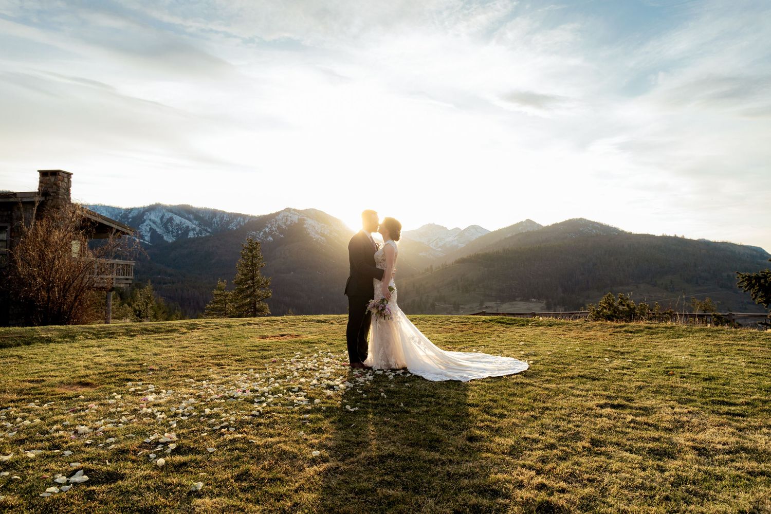 Couple embraces on a scenic mountain meadow during golden sunset with dramatic mountain ranges in the background.