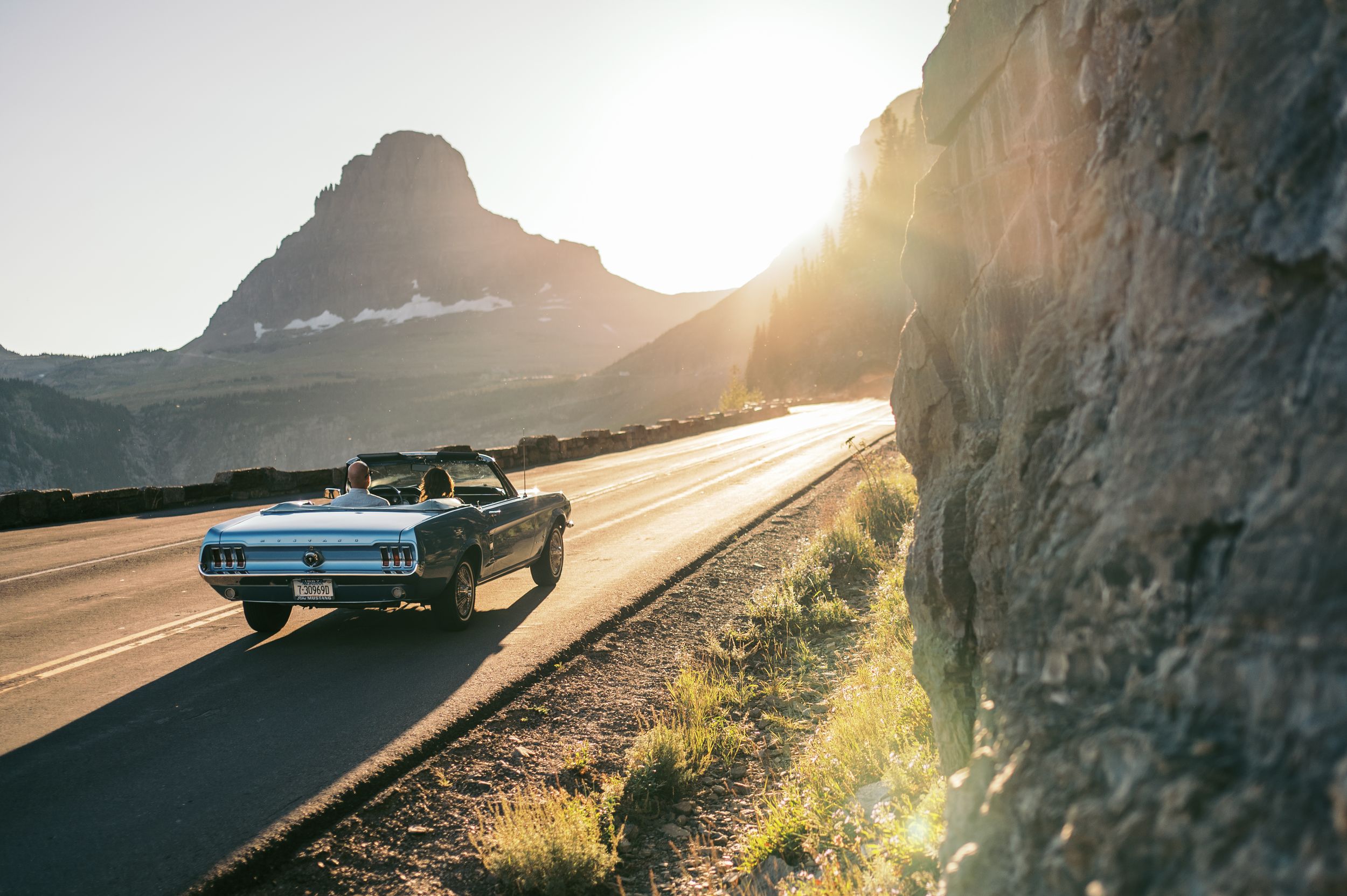 Classic blue car drives along scenic mountain road at golden sunset with dramatic cliffside views.