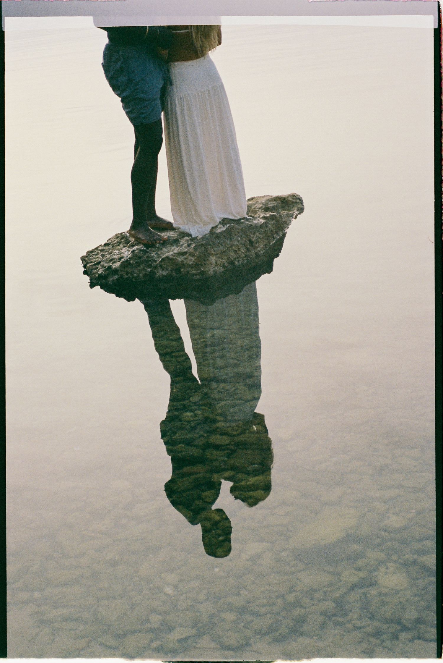 Two people's reflections stand mirrored on a still water surface creating an ethereal double image effect.