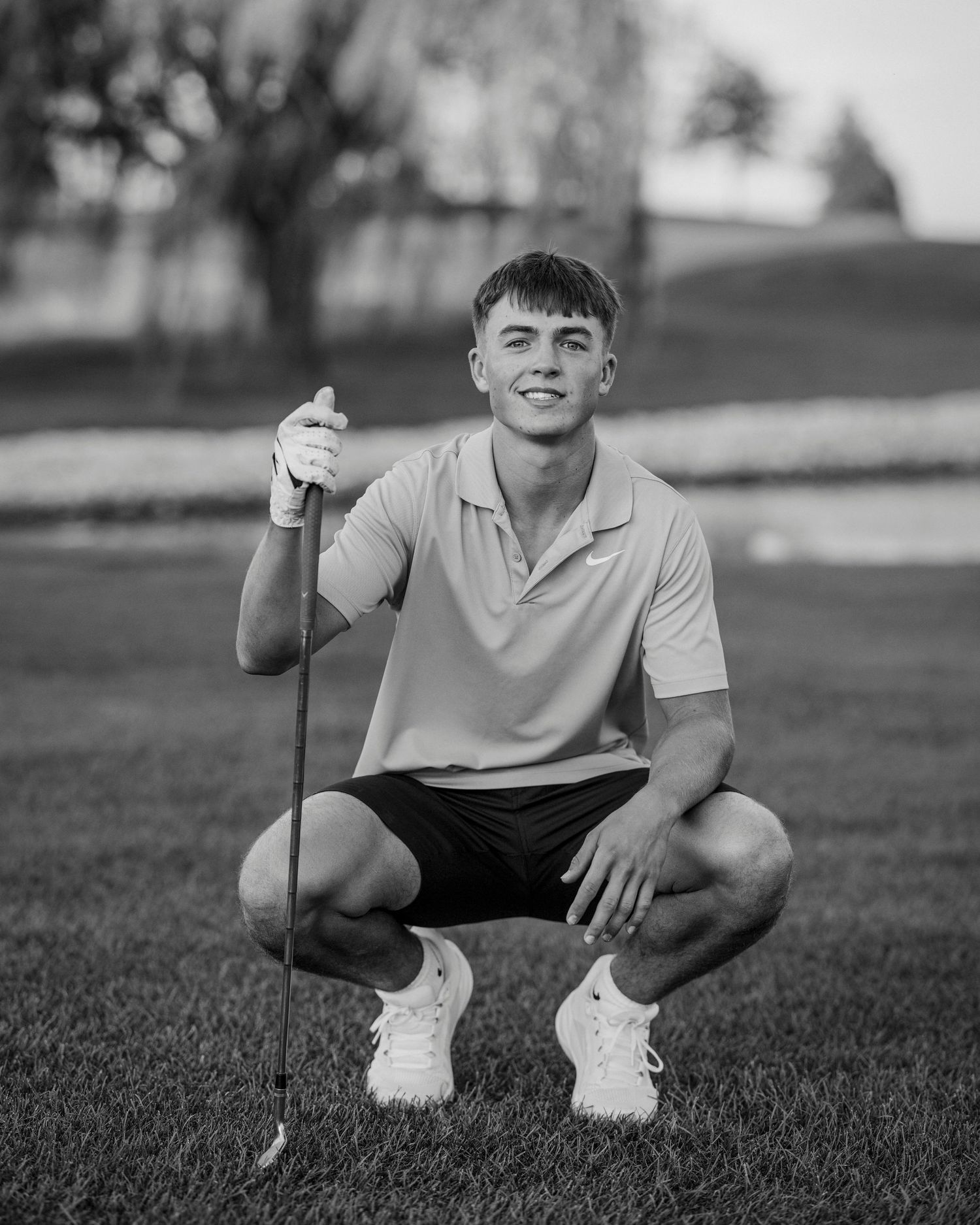 Black and white photo of a young golfer crouching on the green while holding a golf club and smiling at the camera.