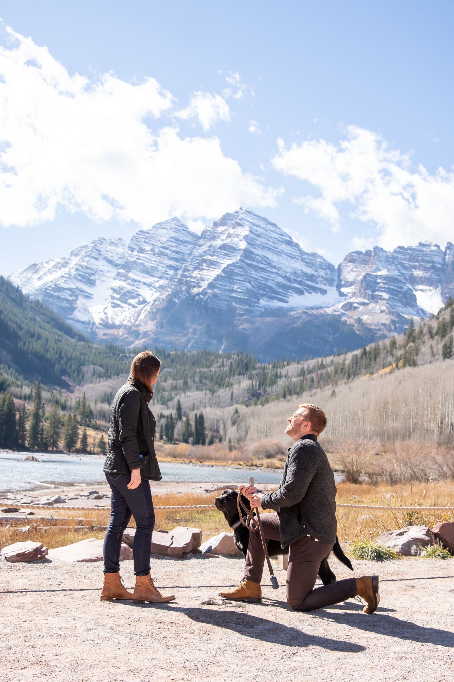 A couple and their dog enjoy a scenic mountain view at a snowy lake with dramatic peaks in the background.