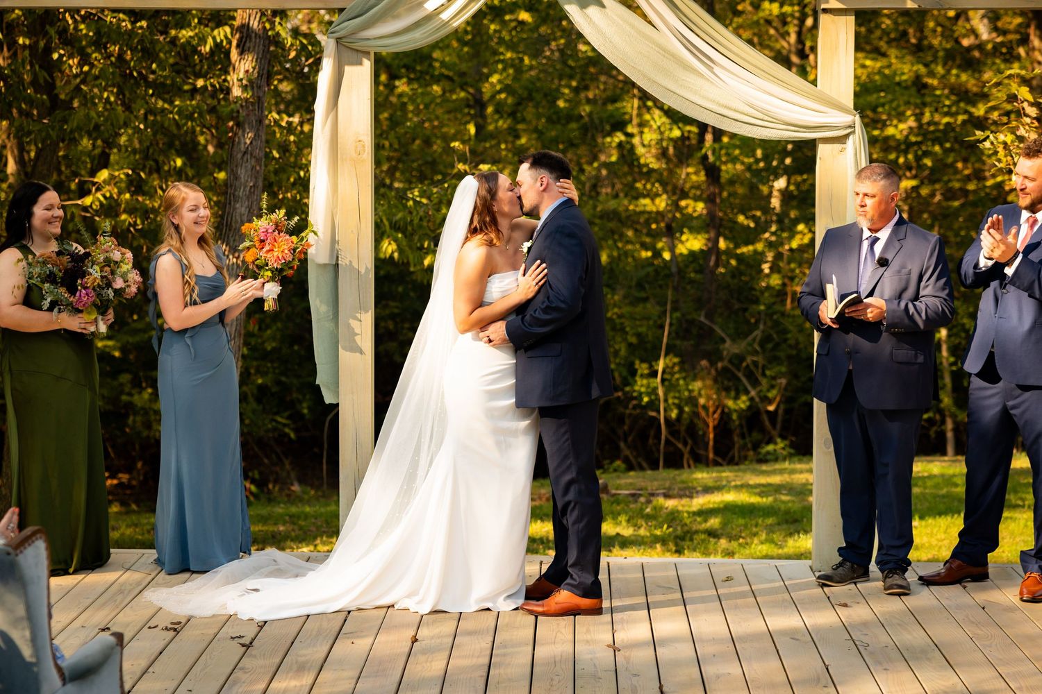 A wedding ceremony takes place on a scenic outdoor deck surrounded by trees and white draping decor.