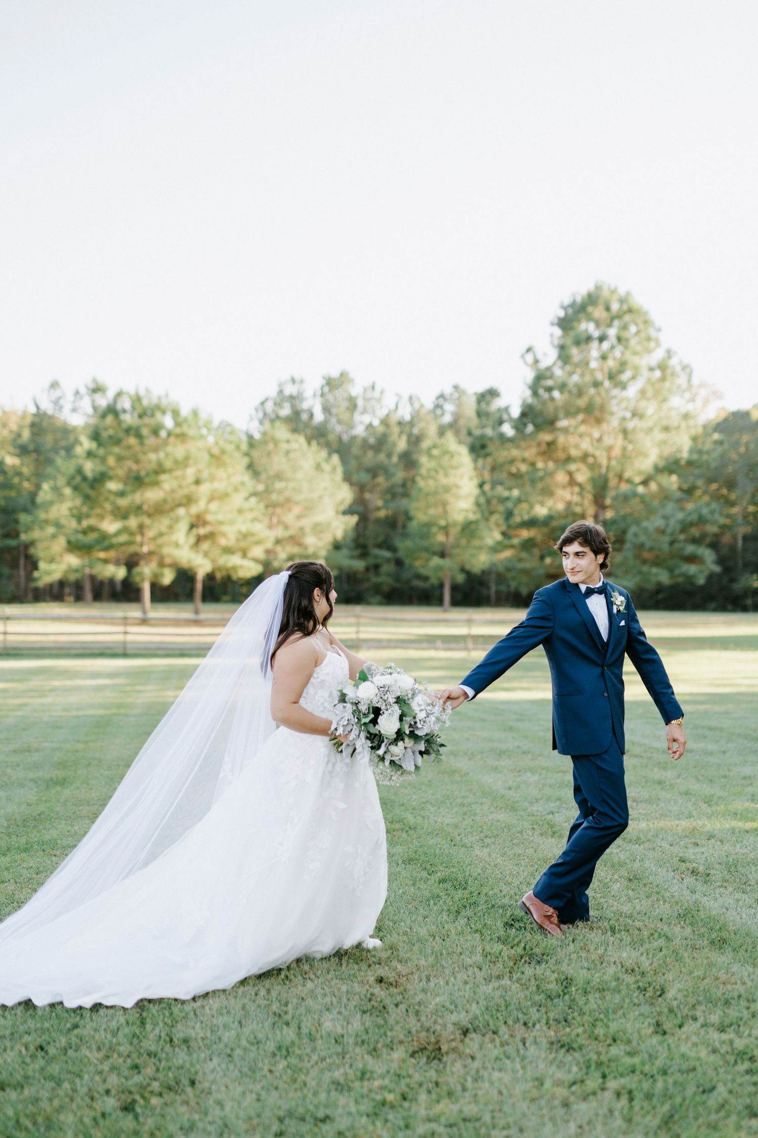 A couple in wedding attire playfully runs across a sunlit grassy field during their outdoor photo session.