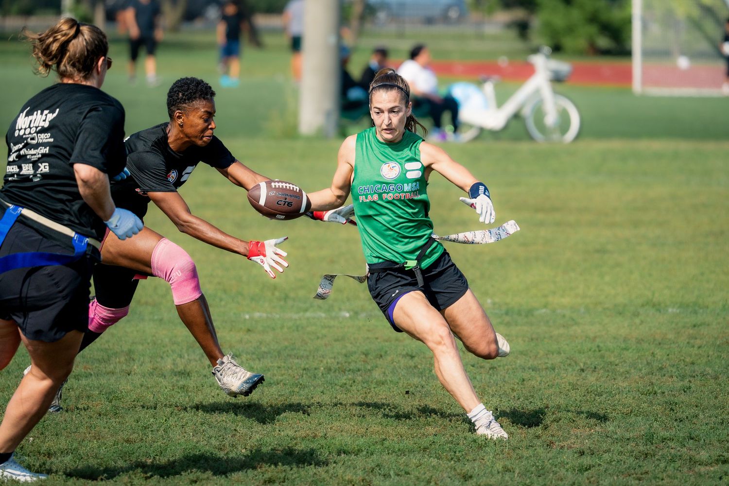 A flag football player in a green jersey with the football runs downfield with the football while an opponent in a black jersey pursues