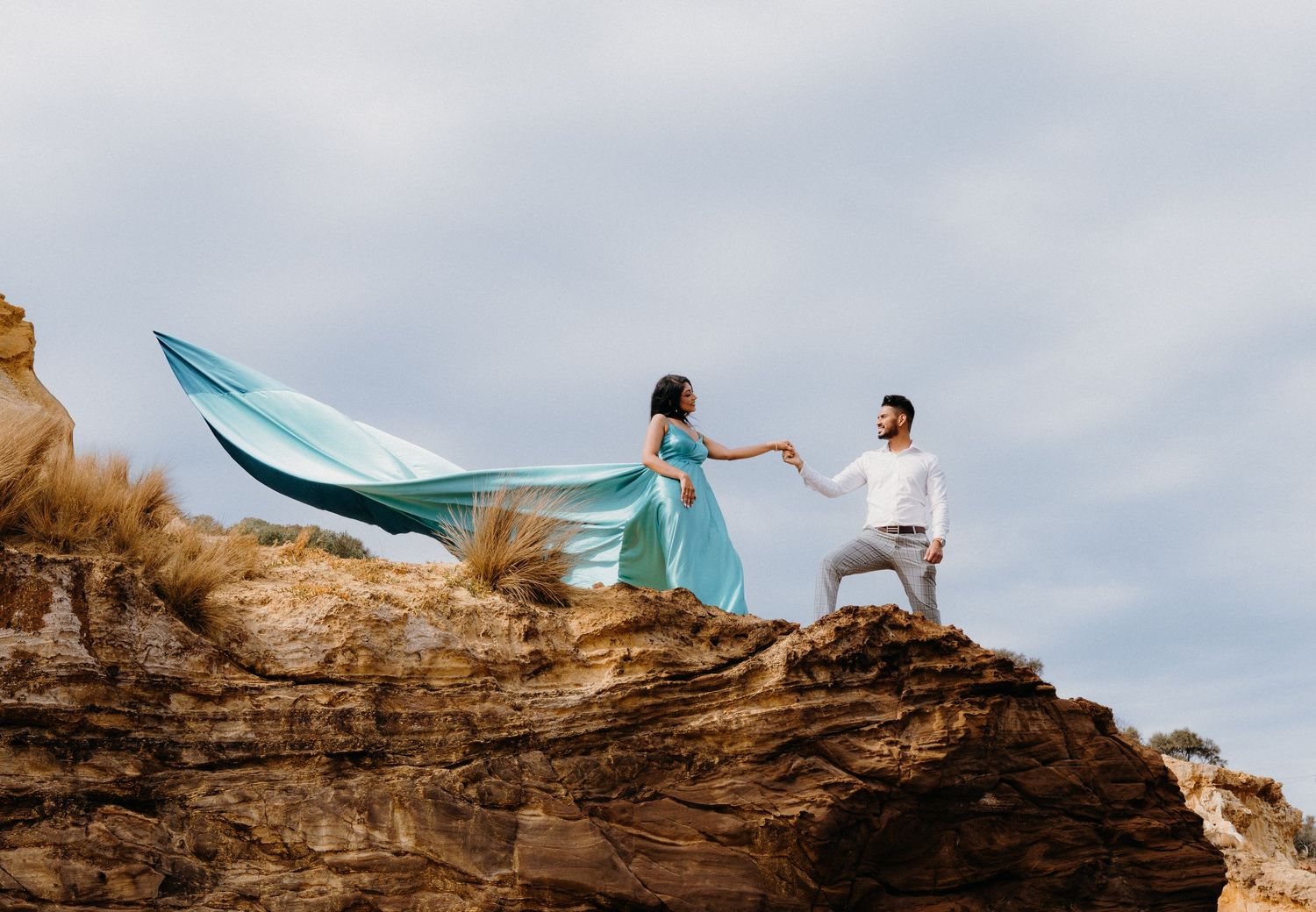Two people in light colored clothing stand on a rocky cliff edge as fabric billows dramatically in the wind.