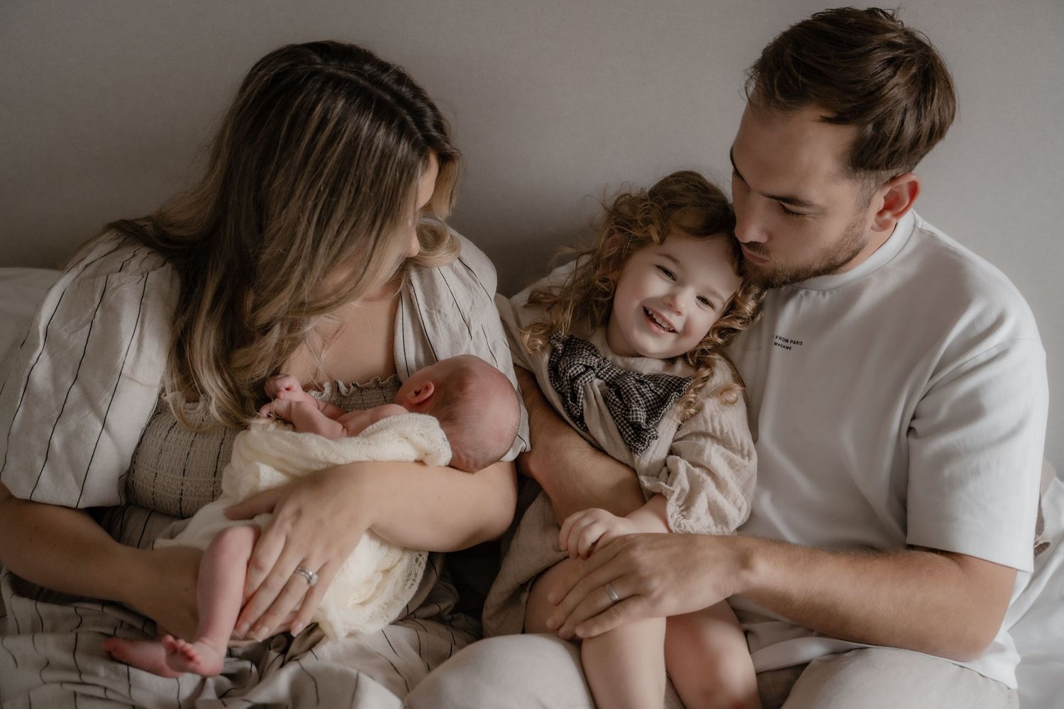 Family sits together on white bed sharing tender moment with newborn baby and toddler.