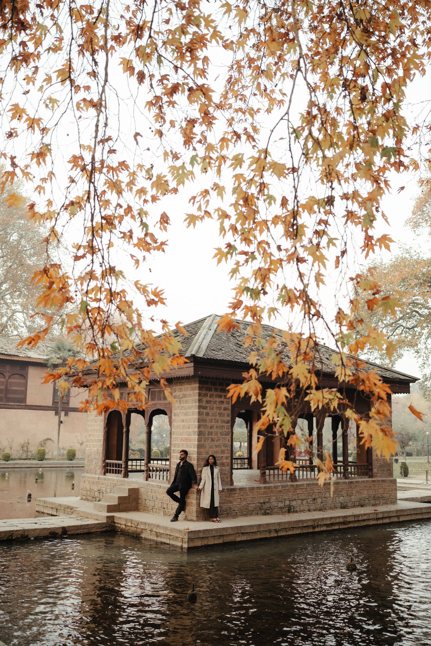 Autumn leaves frame a tranquil stone building with arched walkways along a peaceful waterfront.