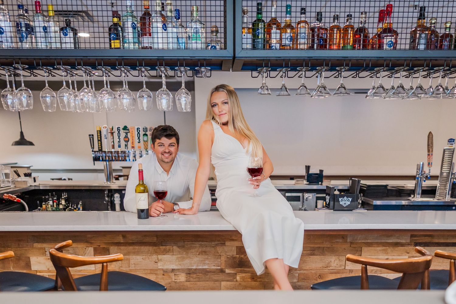 A couple enjoying drinks at a modern bar counter with wine glasses and bottle displays on shelves above.
