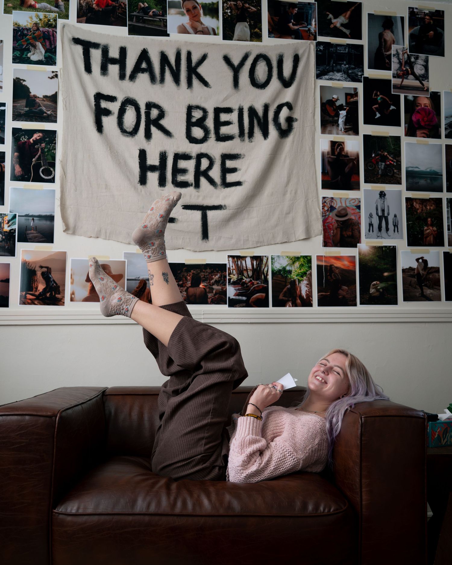 A wall decorated with photographs displays a banner reading 'THANK YOU FOR BEING HERE' above a person resting on a couch.