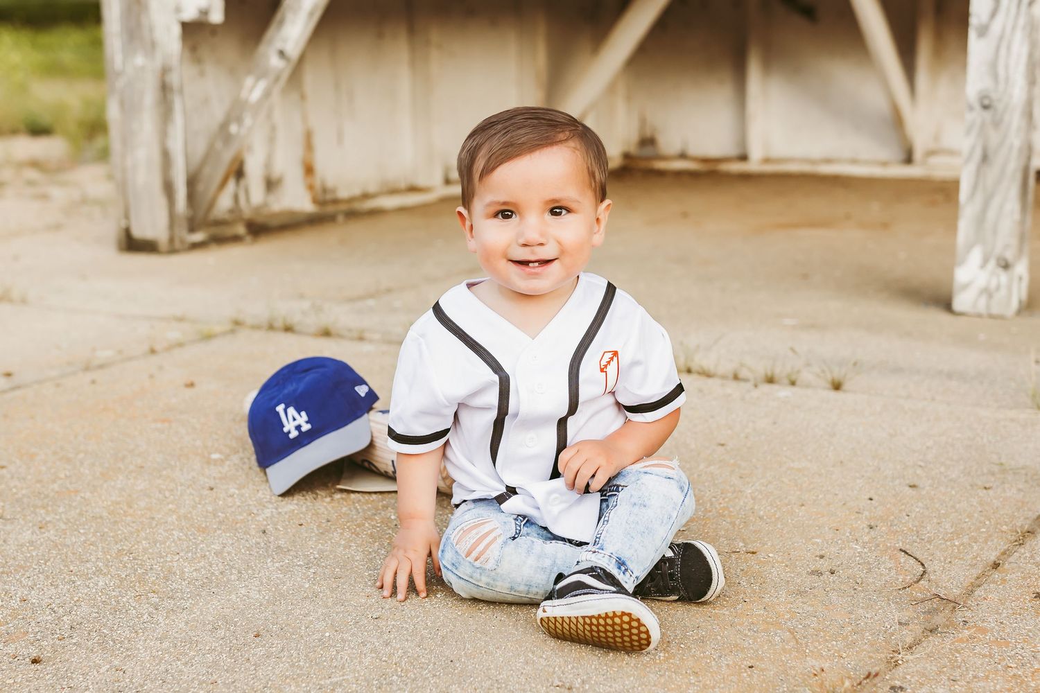 Child in blue baseball jersey and striped pants sits on sandy ground while playing outdoors.