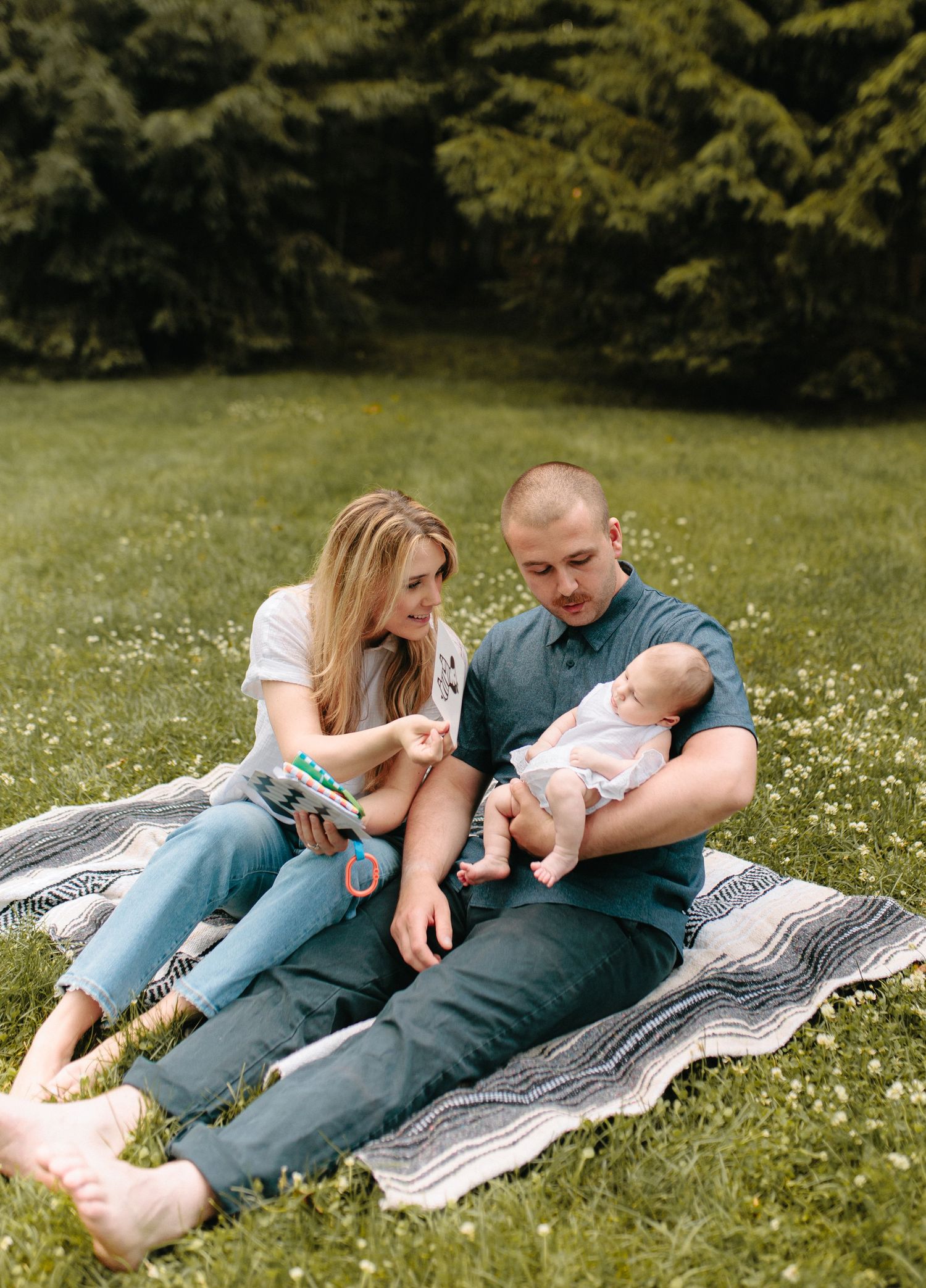 A couple and infant relax on a blanket in a grassy field with wildflowers, surrounded by trees on a sunny day.