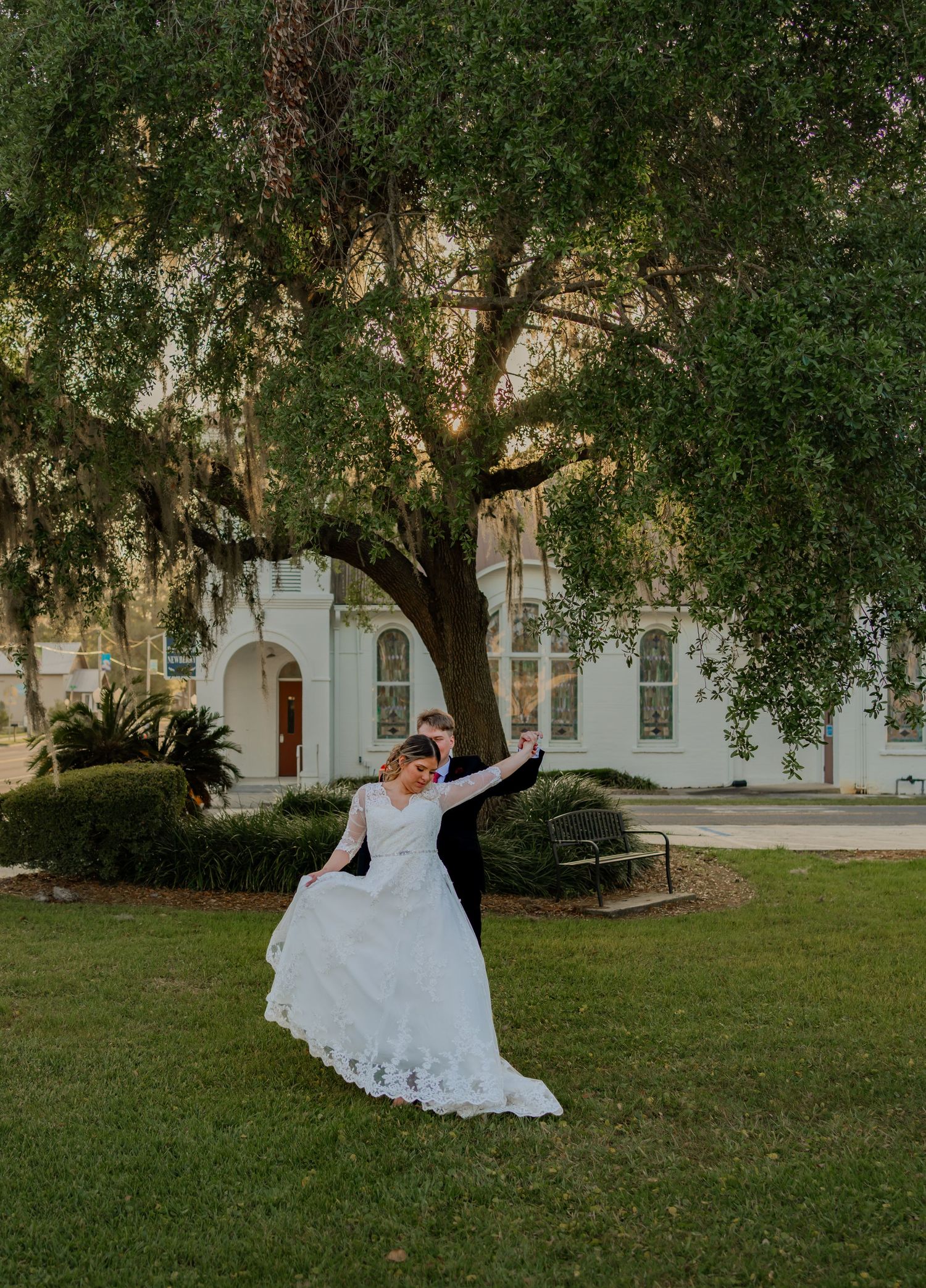 A bride in a flowing white lace dress twirls on a lawn in front of a grand white building under a majestic oak tree.