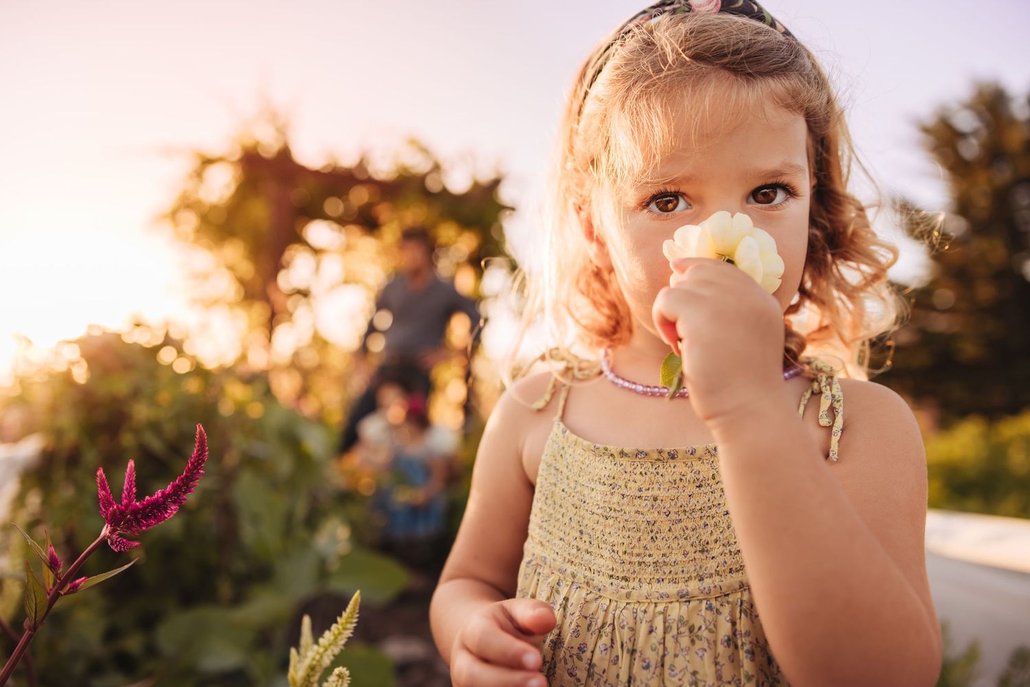 Young girl in a knitted sundress smells a pale yellow flower in a sunlit garden with blurred people and greenery behind her.