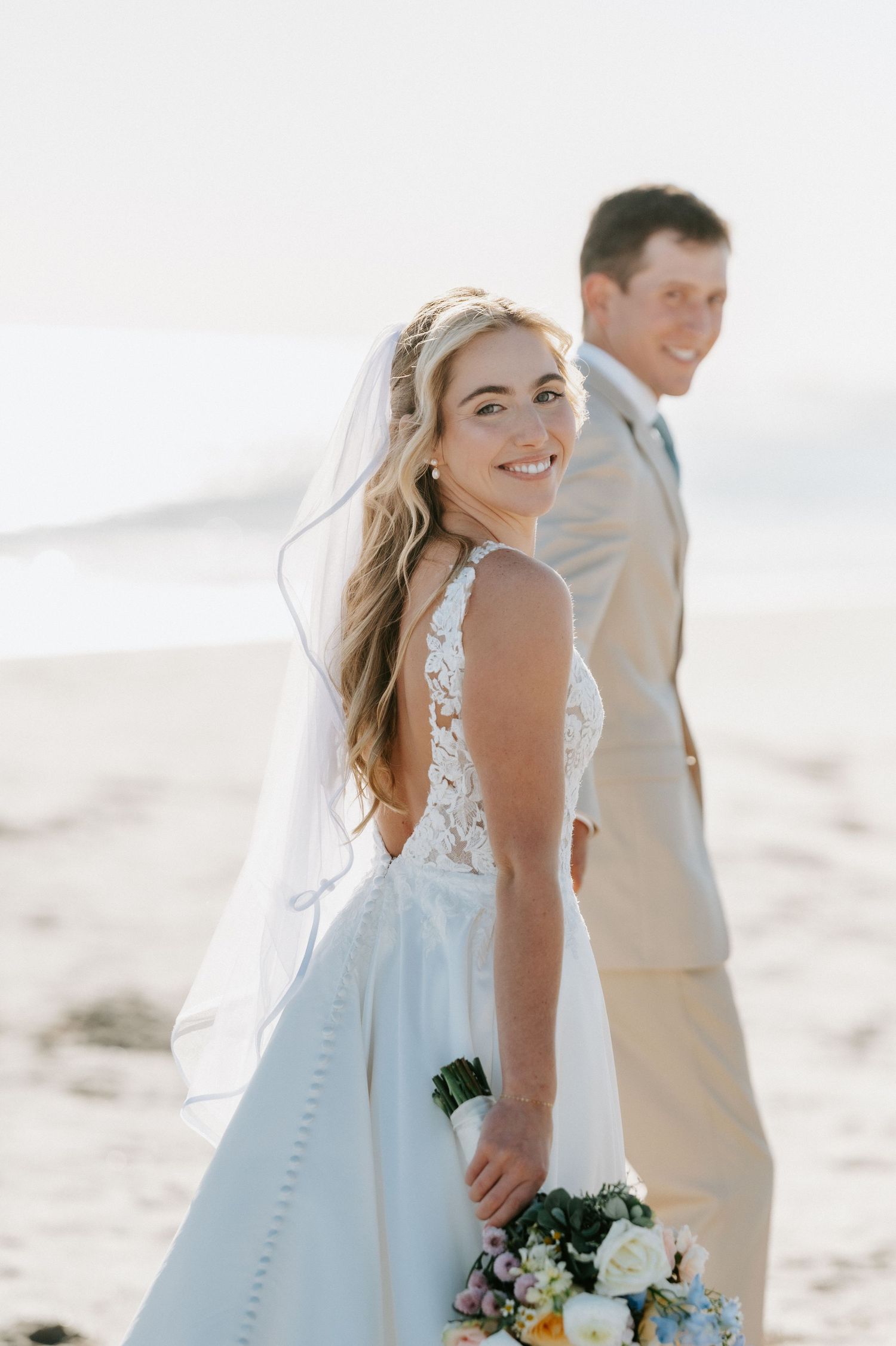 Smiling bride in a lace wedding gown and veil holds a bouquet while walking hand in hand with a groom in a tan suit on a sunny beach.