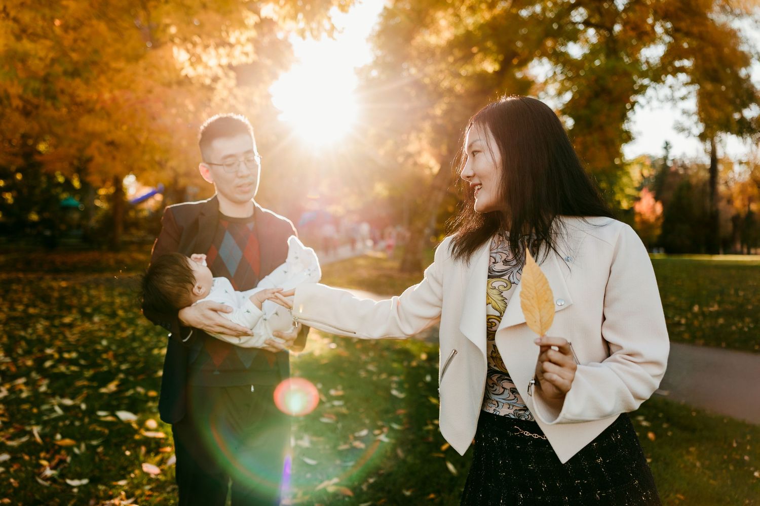 Couple enjoying autumn park with golden sunlight, woman holding yellow leaf, man carrying baby outdoors.