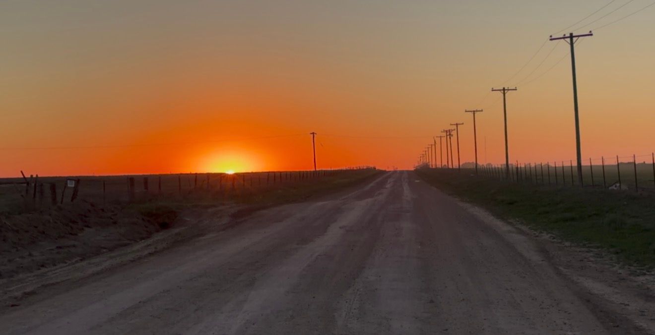 A stunning orange sunset glows over a flat rural dirt road lined with telephone poles stretching to the horizon.