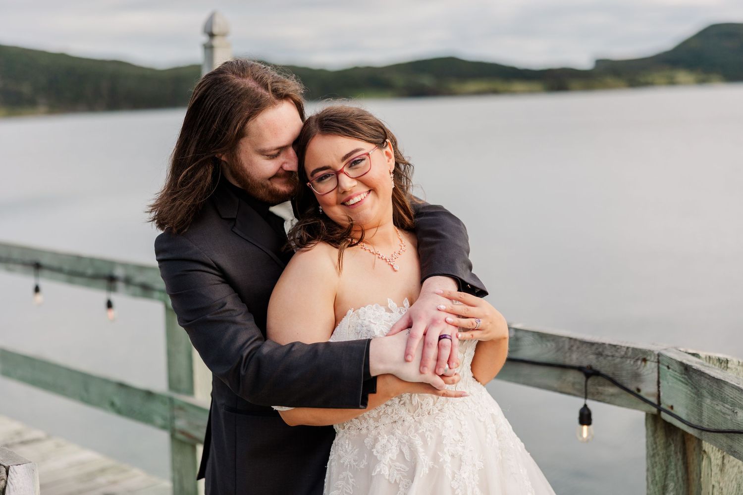 Couple embracing on a lakeside dock, bride in lace gown and groom in black suit, smiling joyfully on their wedding day.