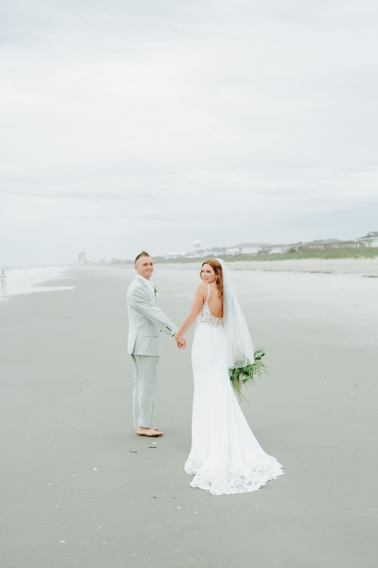Bride and groom hold hands on a beach, she in a white gown with veil, he in a light gray suit.