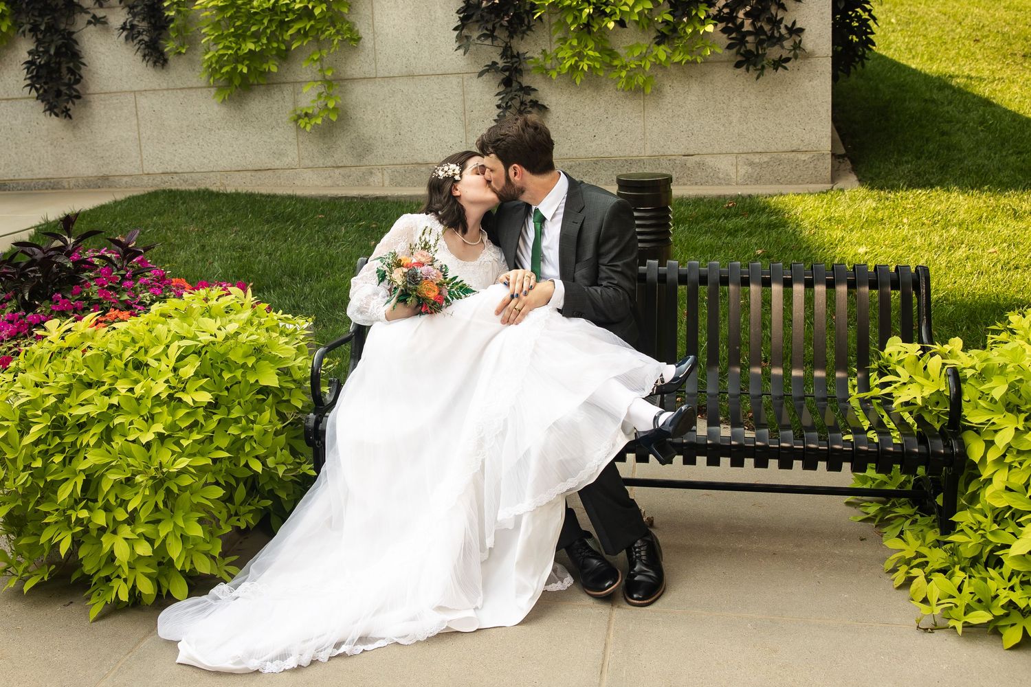 Bride and groom share a kiss outdoors, bride in lace long-sleeve gown, groom in dark suit with green tie.
