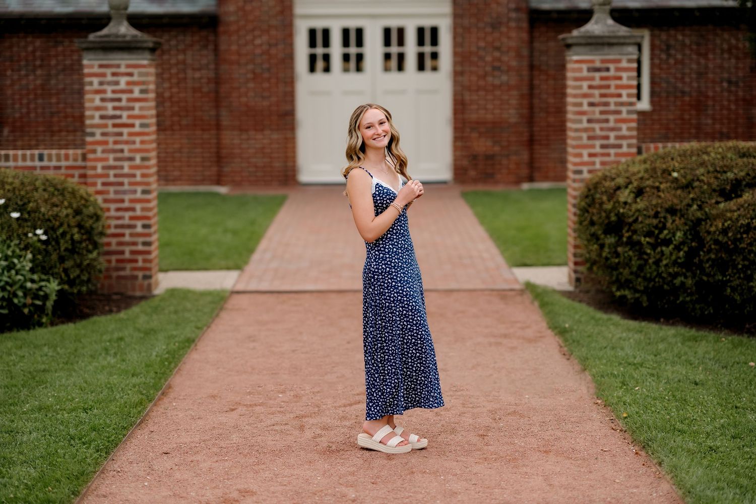 Young woman in navy floral maxi dress smiling on brick path outside historic red brick building.