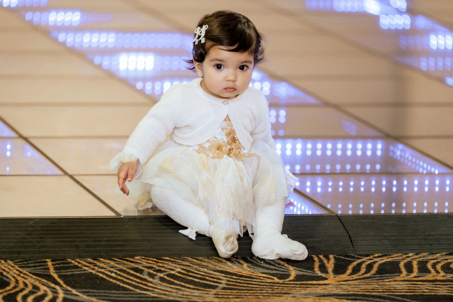 A cute baby girl in a white and gold dress with a hair bow sits on a wooden surface with blue LED lights behind her.