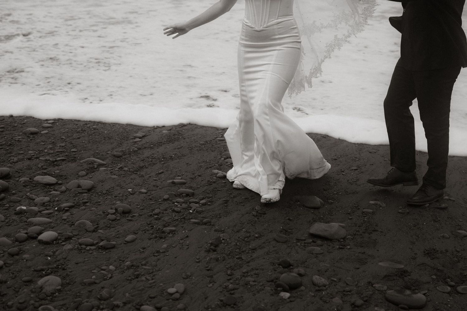 Black and white photo of a bride in a flowing gown walking on a rocky black sand beach near ocean waves.