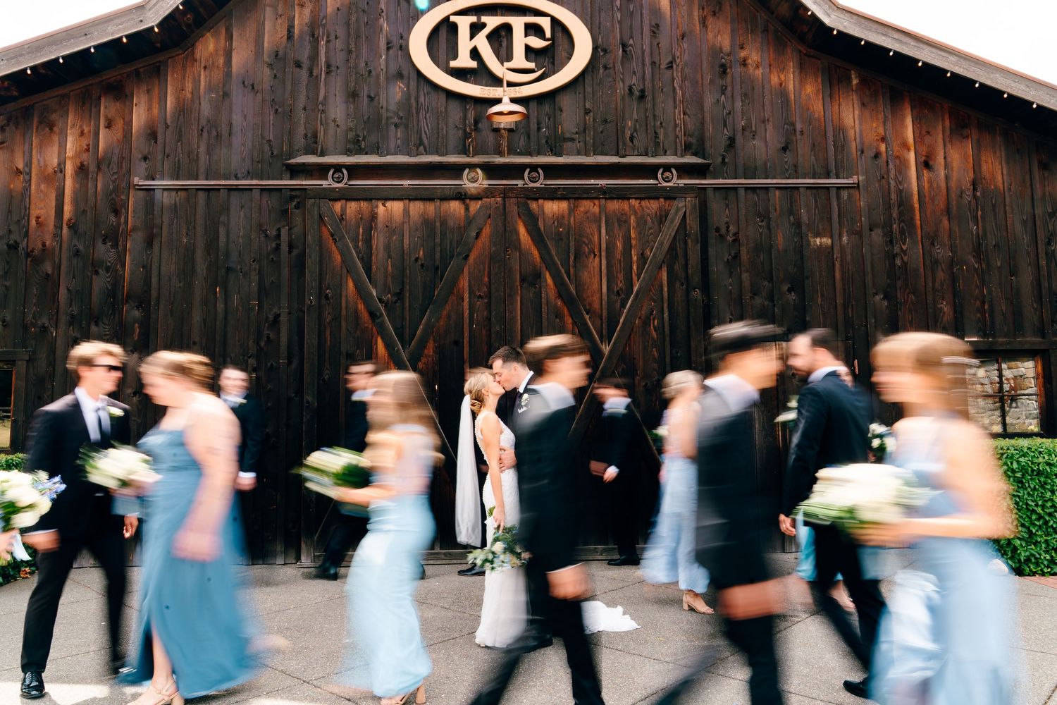 Wedding party walks past rustic barn with KF monogram sign as bride and groom share a kiss.