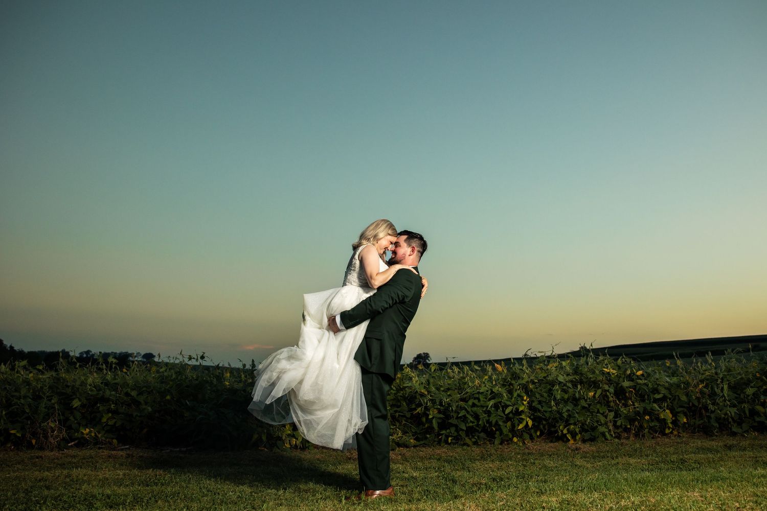 Groom lifting bride in white gown at sunset, sharing a romantic kiss outdoors near a vineyard.
