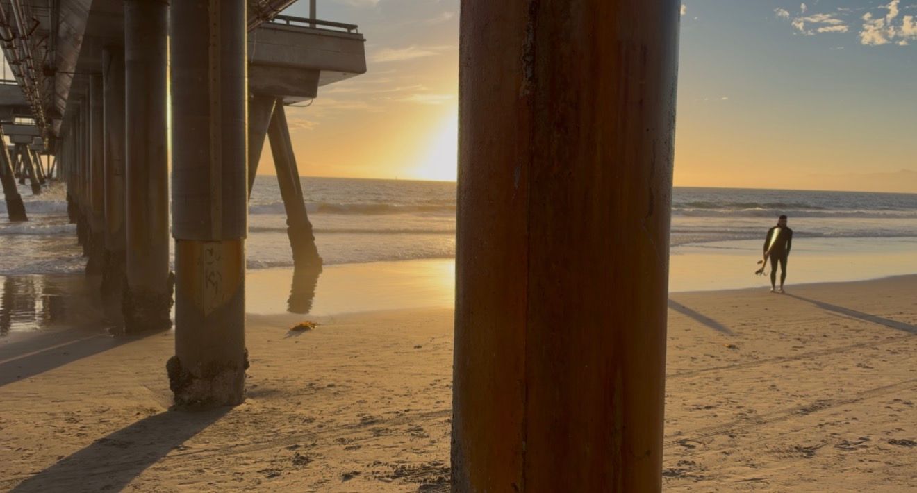 Surfer walks on sandy beach at golden sunset beneath rustic pier pilings in warm evening light.