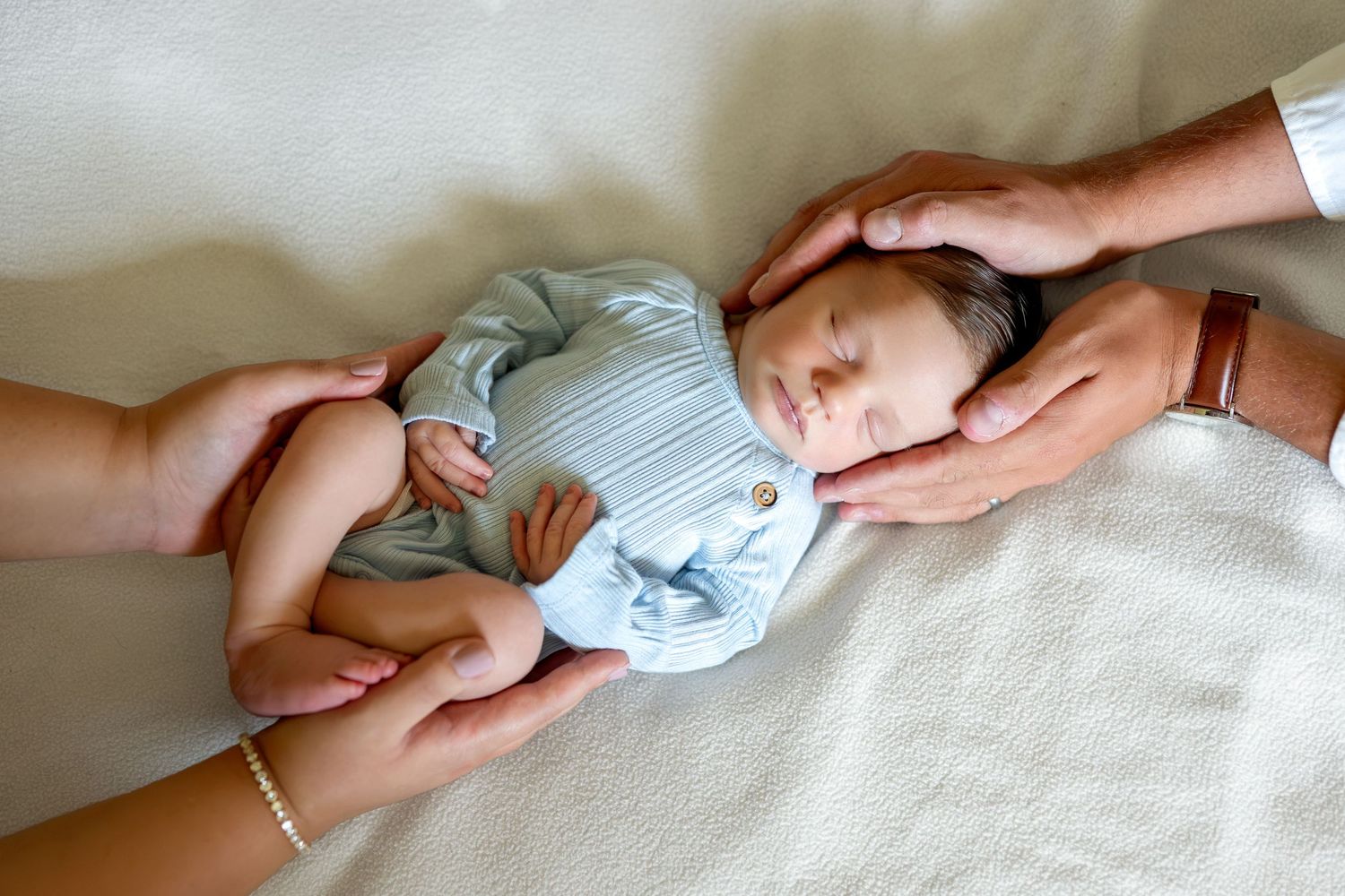 Newborn baby sleeping peacefully in light blue onesie, cradled gently by parents hands on soft white blanket.