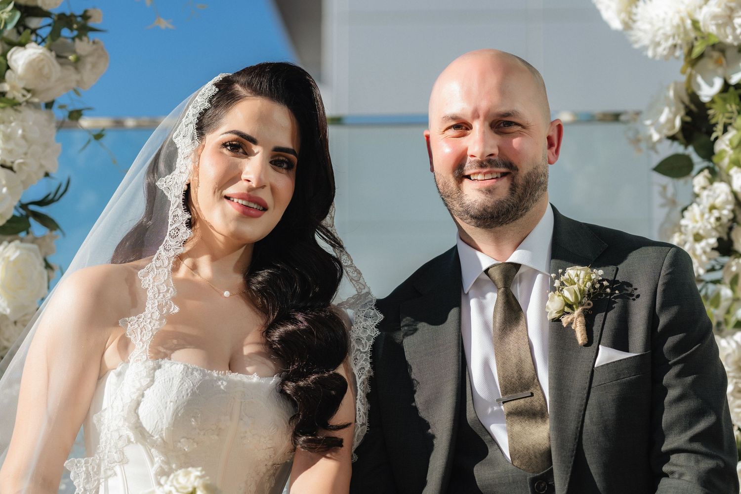 Smiling bride in a lace veil and strapless wedding gown stands beside her bald, bearded groom in a dark suit with a boutonniere among white