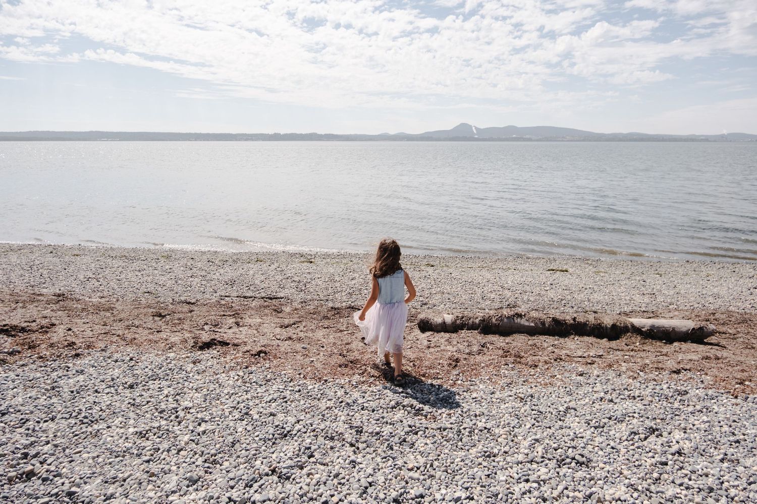 A young girl in a white dress stands on a rocky beach shore, gazing at the calm sea with mountains in the distance.