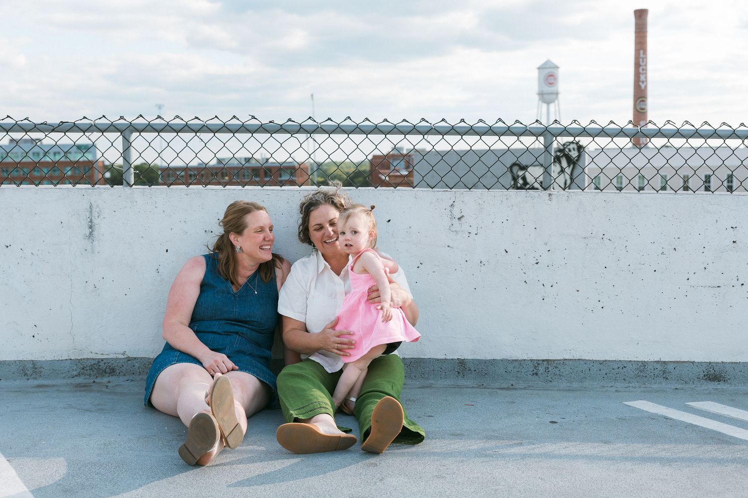 Two women and a toddler sitting together on a rooftop parking deck, smiling and laughing outdoors.