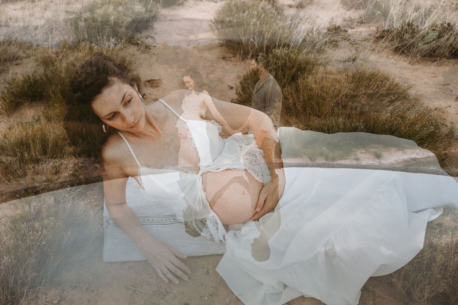 Pregnant woman in white dress holds hands with man by reflective pool in desert landscape.