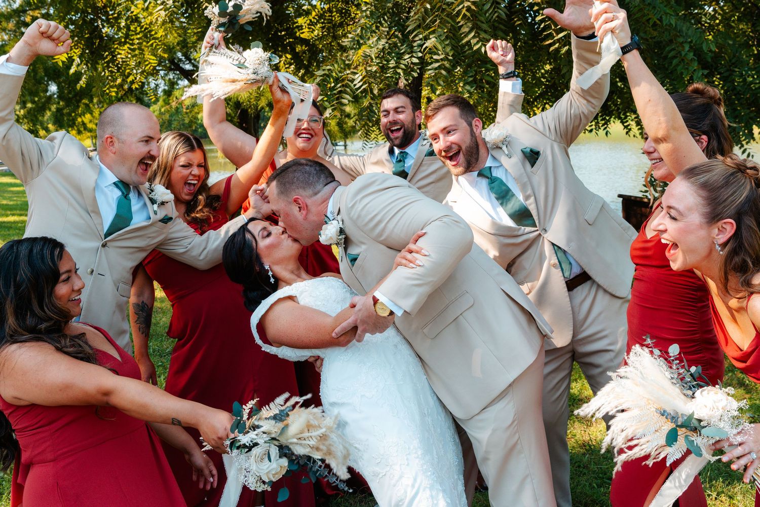 Wedding party celebrates as bride and groom share a kiss, surrounded by cheering guests in red and tan attire outdoors.