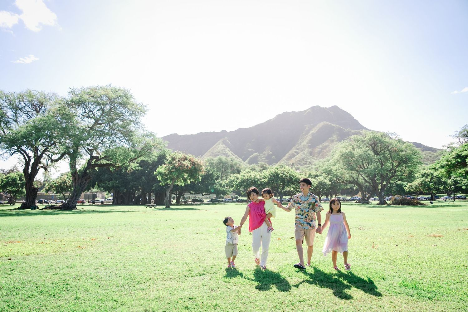 Family of four holding hands walking on green grass with Diamond Head crater and trees in background on sunny day.