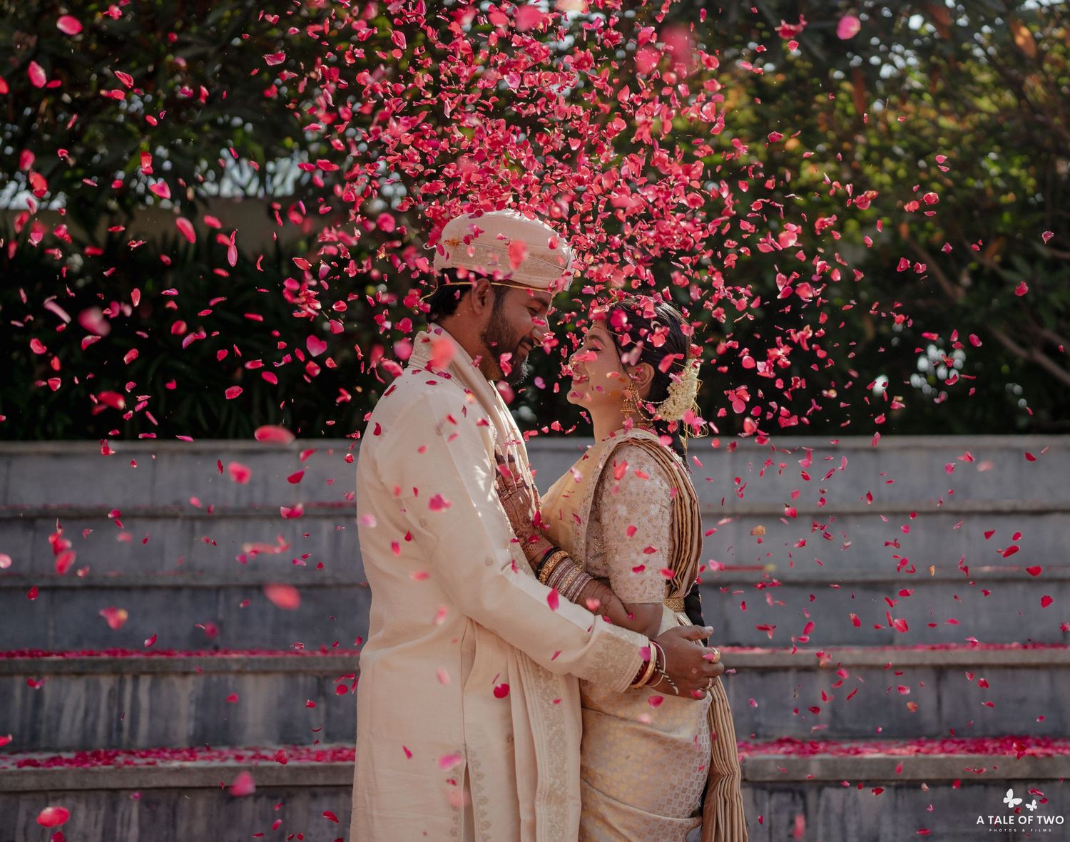Pink rose petals shower a couple in traditional South Asian wedding attire as they share a romantic moment on stone steps.