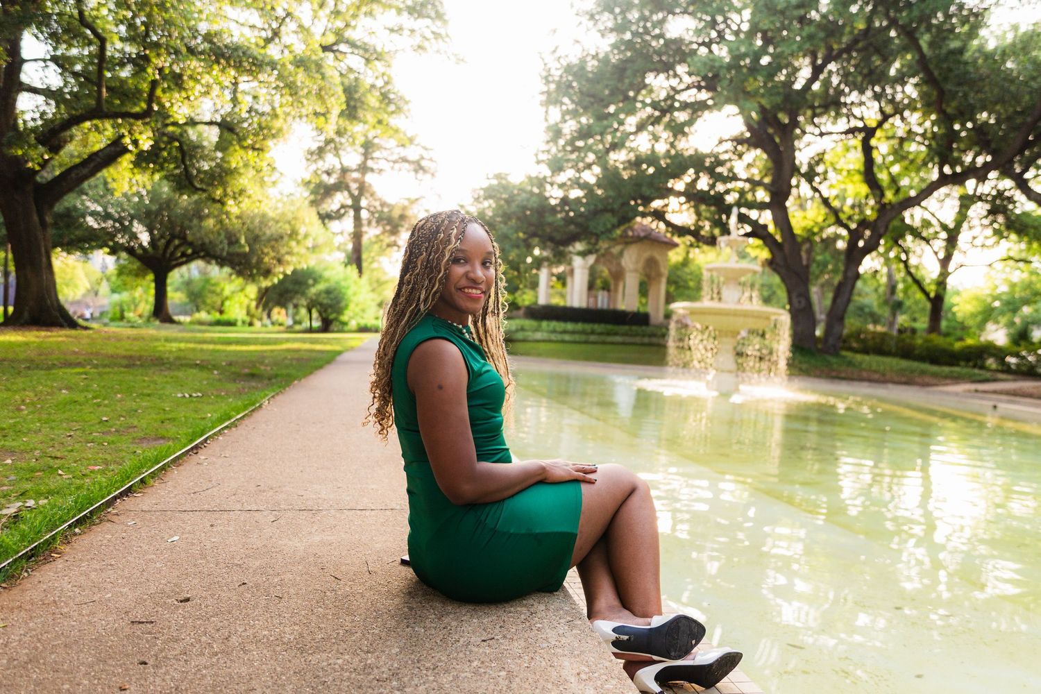 A young woman in a green dress sits beside a fountain in a lush park, smiling over her shoulder.