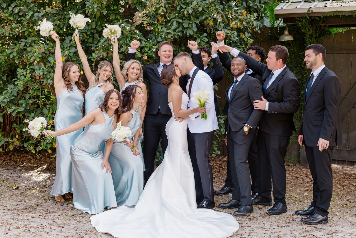 Wedding party celebrating as bride and groom kiss, bridesmaids in blue gowns and groomsmen in black suits outdoors.