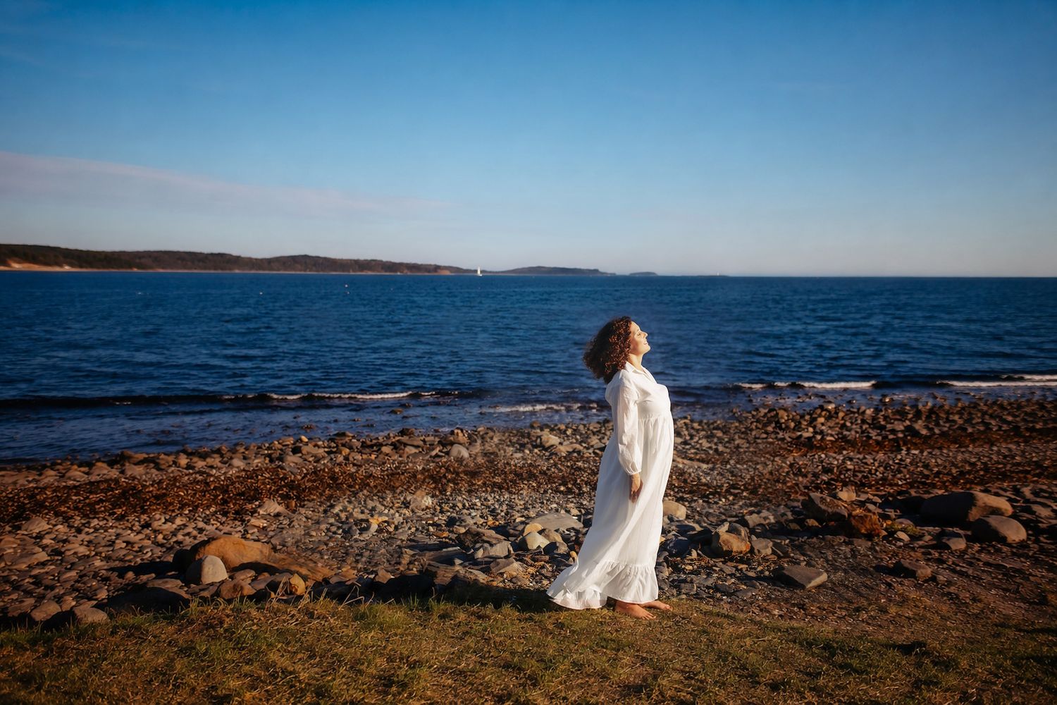 Halifax ns Woman in white maternity session, dress standing on rocky shoreline gazing at calm blue ocean under clear sky.