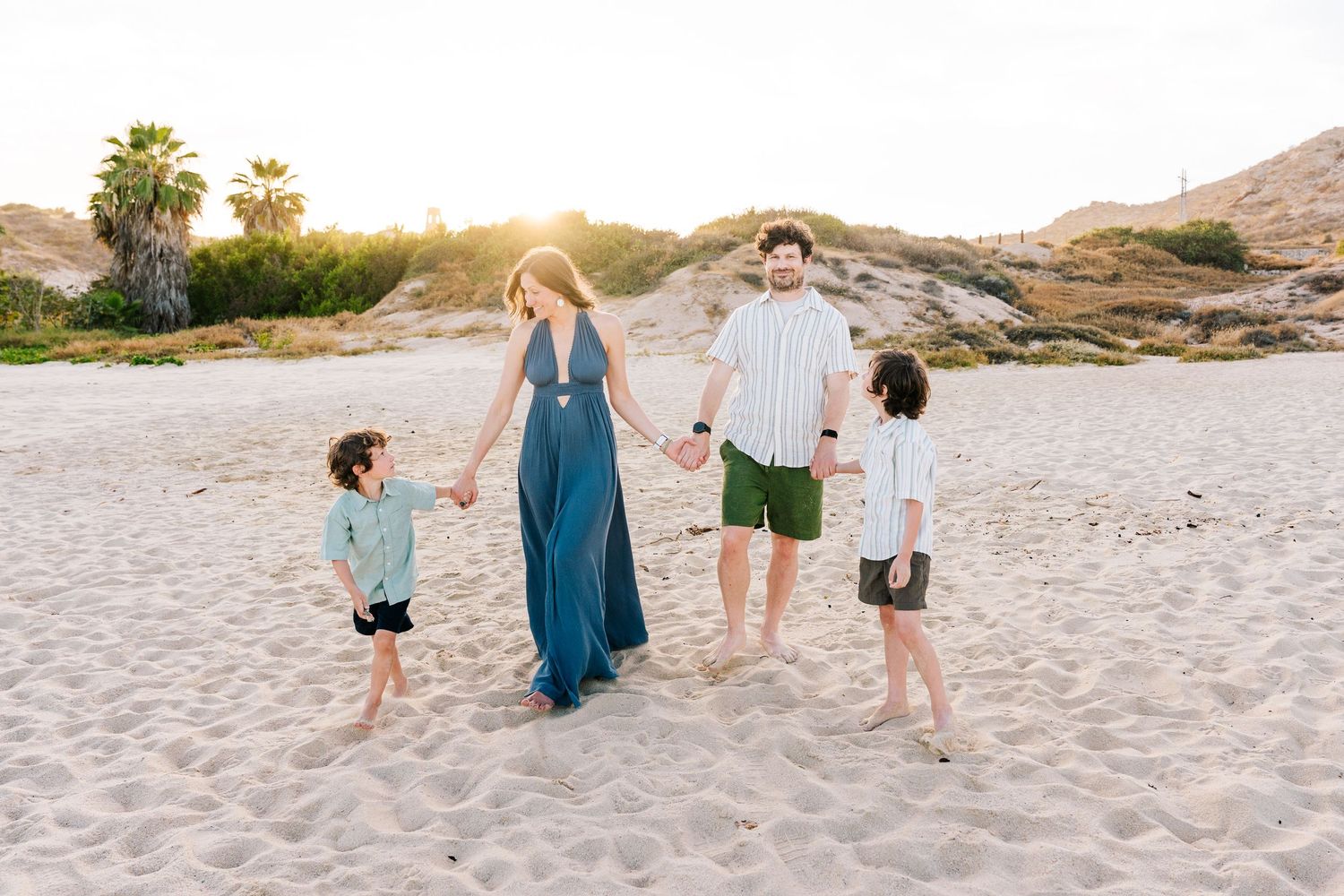 Family of four smiling on sandy beach at golden hour, parents holding young children with warm sunset light behind them.